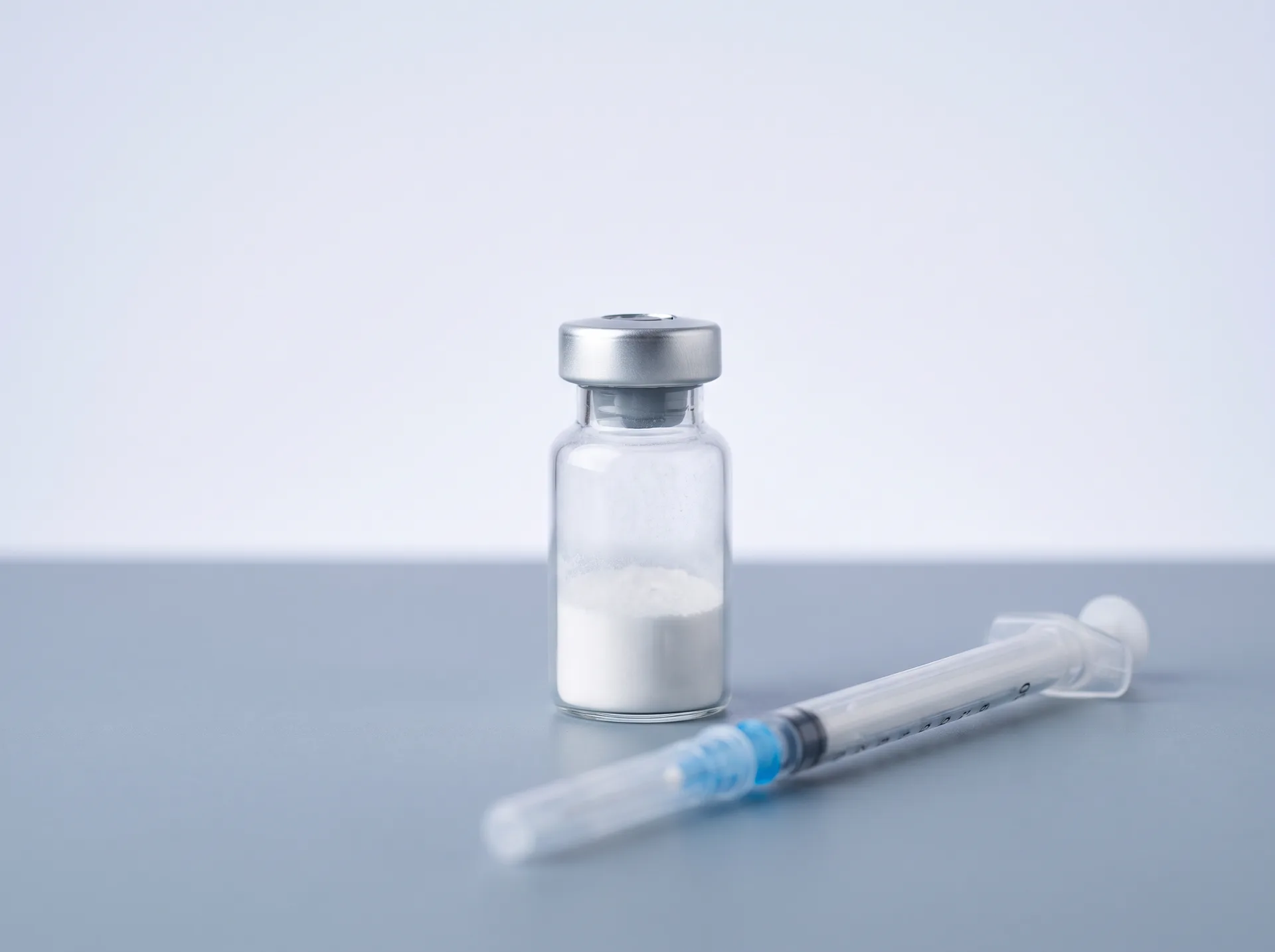 A sealed glass peptide vial beside an insulin syringe on a clean grey surface